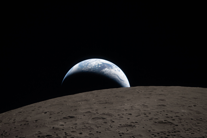 La surface incurvée de la lune au premier plan ressemble à un brun riche, et non à son gris plus familier. Un croissant de terre bleu et blanc s’élève au loin.
