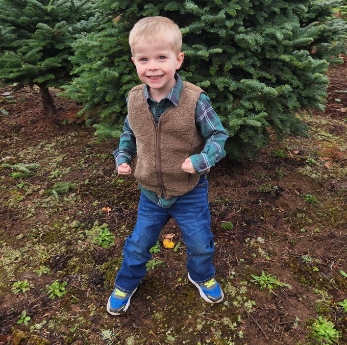 Un jeune garçon souriant aux cheveux blonds se tient dehors, devant un arbre.