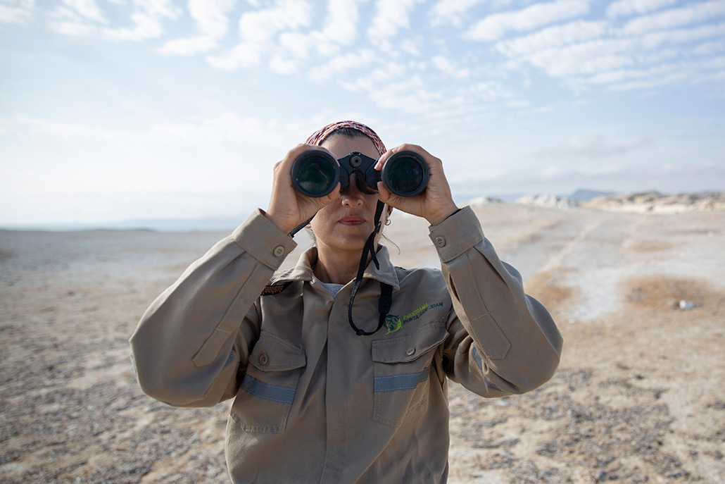 Une vue frontale de Cárdenas-Alayza regardant à travers une paire de jumelles. La côte rocheuse derrière elle s'estompe à mesure qu'elle s'étend au loin.