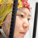 Un participant à l'étude est tourné vers la droite, portant un casque d'électroencéphalogramme (EEG) jaune et rouge qui s'ajuste autour de la tête du participant comme un bonnet de bain. Le casque est doté d'électrodes et d'une bande de puce qui entoure le bas du visage du participant.