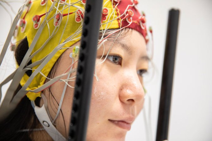 Un participant à l'étude est tourné vers la droite, portant un casque d'électroencéphalogramme (EEG) jaune et rouge qui s'ajuste autour de la tête du participant comme un bonnet de bain. Le casque est doté d'électrodes et d'une bande de puce qui entoure le bas du visage du participant.