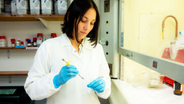 Andrea Gomez porte une blouse de laboratoire et des gants en nitrile bleus et se tient devant une hotte aspirante. Elle regarde un tube à essai qu'elle tient de la main gauche. Sa main droite tient ce qui semble être une pipette.
