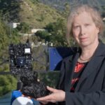 Une femme blanche plus âgée vêtue d'un blazer noir se tenant devant un modèle de vaisseau spatial et tenant un modèle d'objet extérieur du système solaire.