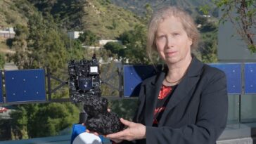 Une femme blanche plus âgée vêtue d'un blazer noir se tenant devant un modèle de vaisseau spatial et tenant un modèle d'objet extérieur du système solaire.