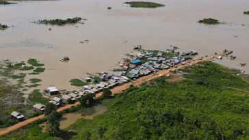 Cette vue aérienne d'une partie du fleuve Sesan, affluent du Mékong, montre une route qui débouche jusqu'au bord du fleuve, bordée de maisons. La rivière est une étendue boueuse parsemée de quelques taches de végétation.