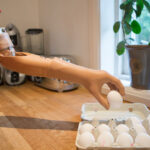 Une personne portant une prothèse de bras bionique ramasse un œuf dans un carton posé sur un comptoir de cuisine.