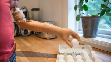 Une personne portant une prothèse de bras bionique ramasse un œuf dans un carton posé sur un comptoir de cuisine.