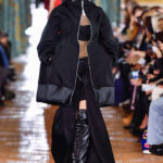 Modèle Hanne Gaby Odiele marche sur une piste avec des cheveux blonds lisses et portant une veste noire surdimensionnée, un haut beige avec une grande bande noire à travers la poitrine, une jupe noire avec une fente de cuisse de cuisse et des bottes en cuir de cuisse noire épais. Odele est intersexué.