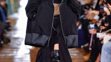 Modèle Hanne Gaby Odiele marche sur une piste avec des cheveux blonds lisses et portant une veste noire surdimensionnée, un haut beige avec une grande bande noire à travers la poitrine, une jupe noire avec une fente de cuisse de cuisse et des bottes en cuir de cuisse noire épais. Odele est intersexué.