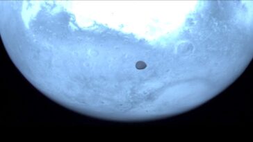 Mars appears light blue in this near-infrared image from the Hera spacecraft, with its moon Deimos represented by a dark mark towards the centre of the image