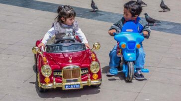 E06NGF An upper class Bolivian girl sits in her very fancy toy remote controlled car that is admired by a poorer boy in Sucre, Bolivia.