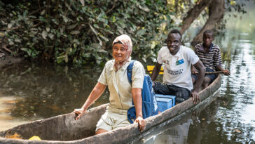 Trois personnes s'assoient dans un bateau en bois flottant sur une rivière. Un refroidisseur bleu se trouve entre la première personne. Ils livrent des vaccins aux personnes dans des endroits éloignés de Sierra Leone.