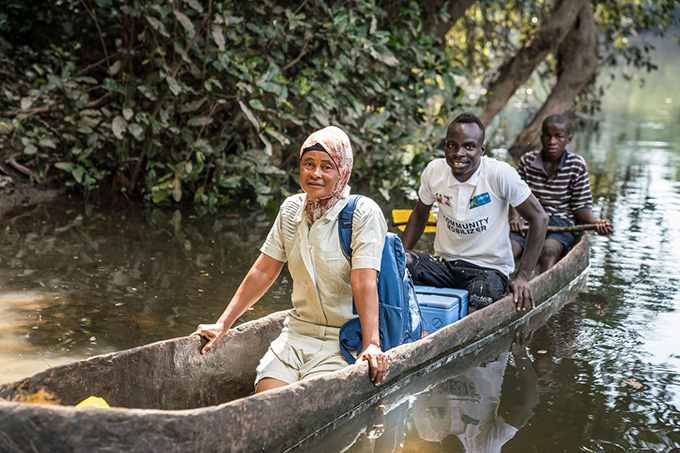 Comment les coupes de santé publique américaines pourraient augmenter les risques de maladies infectieuses Trois personnes s'assoient dans un bateau en bois flottant sur une rivière. Un refroidisseur bleu se trouve entre la première personne. Ils livrent des vaccins aux personnes dans des endroits éloignés de Sierra Leone.