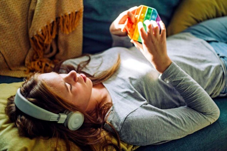 Teenage girl listening to music and playing with push pop toy while lying down on sofa at home