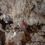 Deux chercheurs portant des casques utilisent des outils pour prélever des échantillons à l'intérieur d'une grotte. L'intérieur est rempli de formations rocheuses comme des stalactites suspendues au plafond de la grotte.
