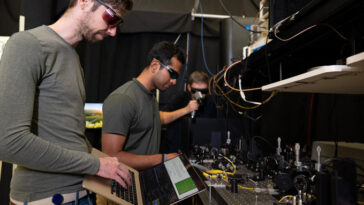 Trois hommes dans un laboratoire, un au premier plan tenant un ordinateur portable ouvert, travaillent sur un projet pour générer des nombres aléatoires.