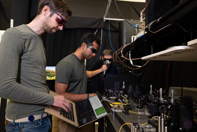 Trois hommes dans un laboratoire, un au premier plan tenant un ordinateur portable ouvert, travaillent sur un projet pour générer des nombres aléatoires.