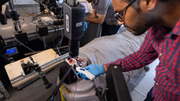 Une personne enveloppée dans un costume spécialisé et recouverte d'une couverture se trouve avec seulement sa tête visible. Il est entouré d'équipement et de deux collègues, pour mesurer sa sueur.