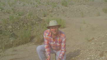 Un chercheur en jean, une chemise à manches longues à carreaux et un soleil s'agenouillent sur un sol sablonneux et rocheux, tenant des outils pour rechercher le sol des fossiles.