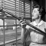 F14FTY 1950s WOMAN VACUUMING VENETIAN BLINDS ON WINDOW LOOKING OUT ONTO SUBURBAN STREET
