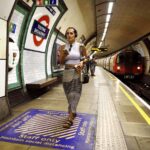 2JHRXY3 London, England, UK. London Underground - young woman with mobile phone and wearing headphone walking along the platform of Tooting Broadway station