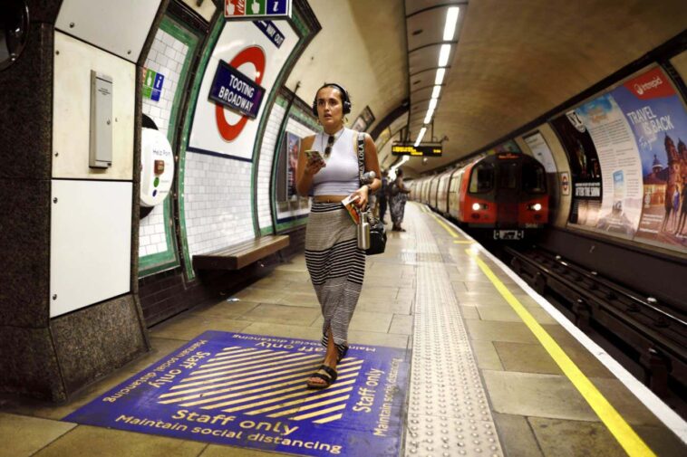2JHRXY3 London, England, UK. London Underground - young woman with mobile phone and wearing headphone walking along the platform of Tooting Broadway station