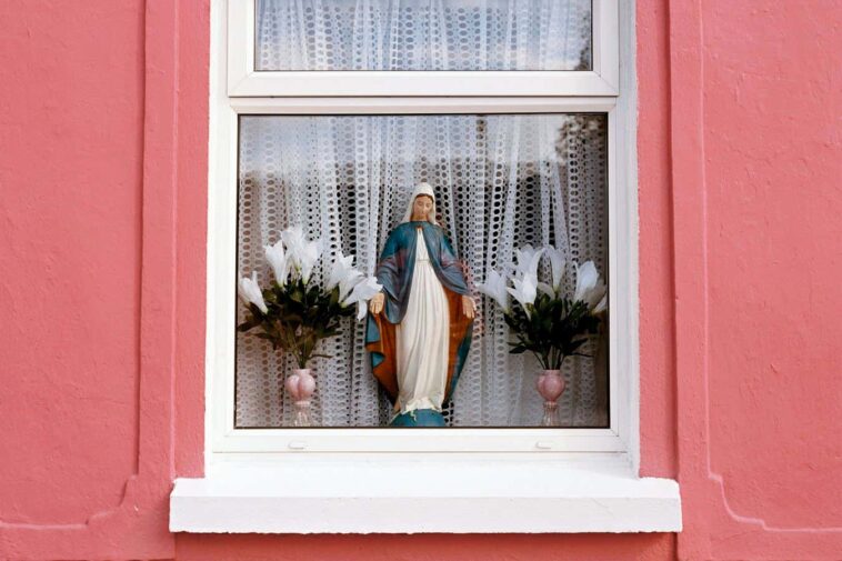 Holy Mary on window Statuette of Holy Mary and vase with plastic flowers in window on a house with bright pink walls, Irish cottage, Ireland
