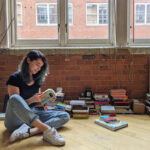 Une femme est assis en tailleur croisée sur le sol en regardant à travers un livre. Des tas d'autres livres sont derrière elle.