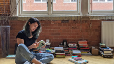 Une femme est assis en tailleur croisée sur le sol en regardant à travers un livre. Des tas d'autres livres sont derrière elle.