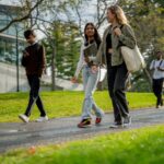 A group of university students are seen walking outside on campus as they make their way from residence to class. They are each dressed casually and have backpacks and book bags with them.
