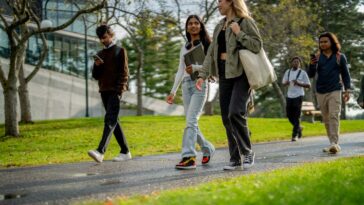 A group of university students are seen walking outside on campus as they make their way from residence to class. They are each dressed casually and have backpacks and book bags with them.