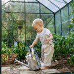 An infant holds a watering can in a greenhouse