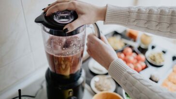 Woman holding blender and making healthy smoothie with various vegetables and fruits in the kitchen at home