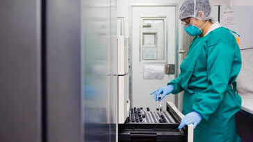 Vue de profil d'un scientifique portant une blouse de laboratoire verte, un masque facial, un filet à cheveux, des gants en latex et des lunettes. Le scientifique tient un tube à essai au-dessus d’un tiroir ouvert à l’intérieur d’un laboratoire.