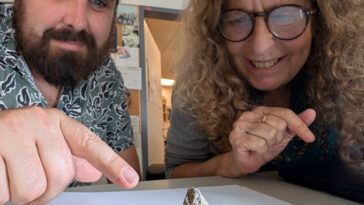 Un homme montre une petite figurine en argile posée sur une feuille de papier blanc sous le regard d’une femme.