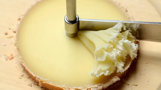 Une meule de fromage est représentée en train d'être grattée pour former une rosette par une lame rotative.