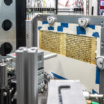 Une page manuscrite fragile est montée dans un cadre métallique à l’intérieur d’un système d’imagerie de laboratoire entouré d’équipements scientifiques.