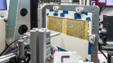 Une page manuscrite fragile est montée dans un cadre métallique à l’intérieur d’un système d’imagerie de laboratoire entouré d’équipements scientifiques.