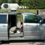 Une fourgonnette Toyota Sienna 2013 argentée avec les portes arrière coulissantes ouvertes. Le fourgon a été modifié avec un équipement cylindrique monté sur le toit. La camionnette est garée sur un parking bétonné devant des arbres et un espace clôturé.