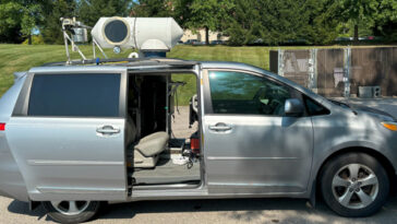 Une fourgonnette Toyota Sienna 2013 argentée avec les portes arrière coulissantes ouvertes. Le fourgon a été modifié avec un équipement cylindrique monté sur le toit. La camionnette est garée sur un parking bétonné devant des arbres et un espace clôturé.