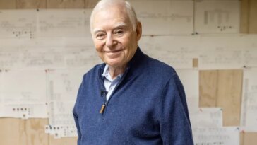 Un homme aux cheveux blancs portant un pull bleu sourit.