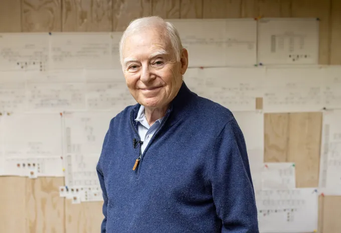 Un homme aux cheveux blancs portant un pull bleu sourit.