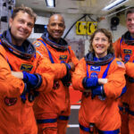 Quatre personnes sont alignées, vêtues de combinaisons spatiales orange.
