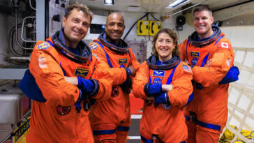 Quatre personnes sont alignées, vêtues de combinaisons spatiales orange.