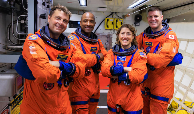 Quatre personnes sont alignées, vêtues de combinaisons spatiales orange.