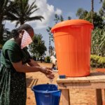 Le Congo se prépare à affronter Ebola. Aujourd’hui, une souche rare révèle des lacunes dans l’état de préparation Une femme portant un foulard sur le visage se lave les mains sur un stand extérieur en tenant de l'eau dans un grand seau orange, avec un seau bleu pour récupérer l'eau usée.
