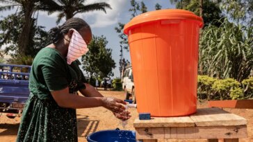Une femme portant un foulard sur le visage se lave les mains sur un stand extérieur en tenant de l'eau dans un grand seau orange, avec un seau bleu pour récupérer l'eau usée.