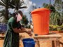 Une femme portant un foulard sur le visage se lave les mains sur un stand extérieur en tenant de l'eau dans un grand seau orange, avec un seau bleu pour récupérer l'eau usée.