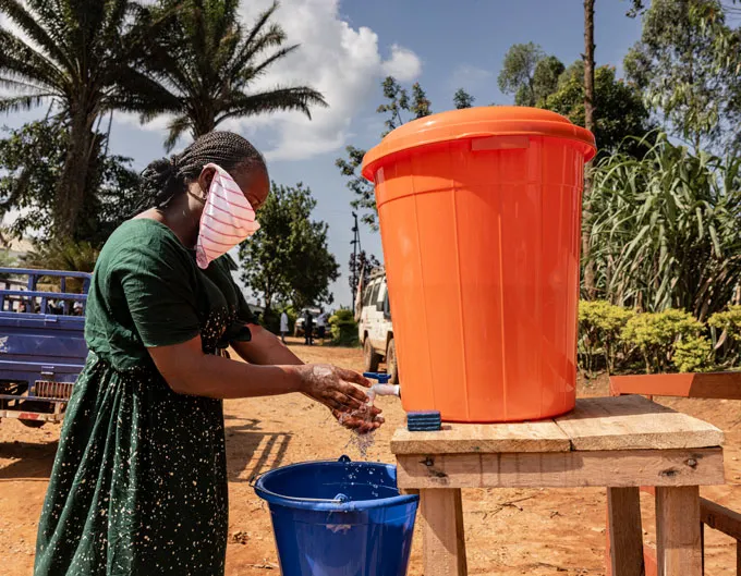 Une femme portant un foulard sur le visage se lave les mains sur un stand extérieur en tenant de l'eau dans un grand seau orange, avec un seau bleu pour récupérer l'eau usée.