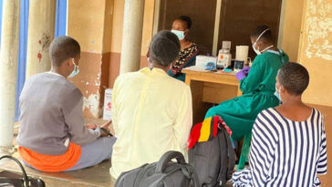 Trois personnes sont assises sur un banc et nous tournent le dos. En arrière-plan, un professionnel de la santé portant une blouse verte, un masque facial et des clous de girofle violets semble préparer un échantillon. L'équipement est posé sur la table où elle est assise et on peut voir une femme assise sur une chaise à côté de la table.